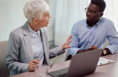 A White Haired Business Woman In Her 60s Conversing With A Black Business Man In His 50s Over A Desk And Laptop