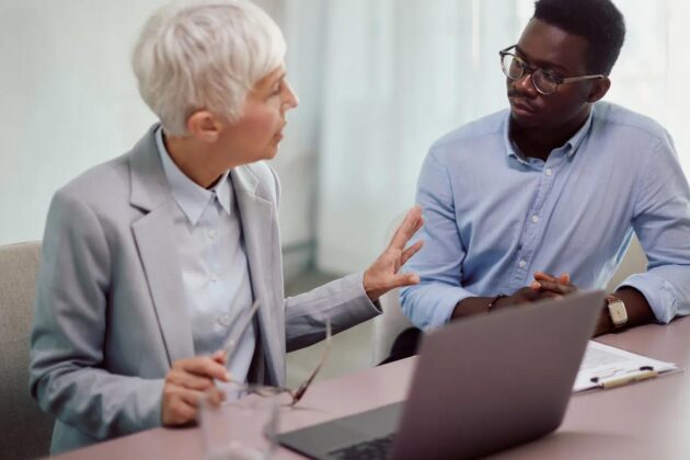 A White Haired Business Woman In Her 60s Conversing With A Black Business Man In His 50s Over A Desk And Laptop