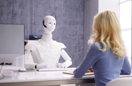 Robot Behind A Desk Conducting A Conversation With A Young Woman In Business Attire.