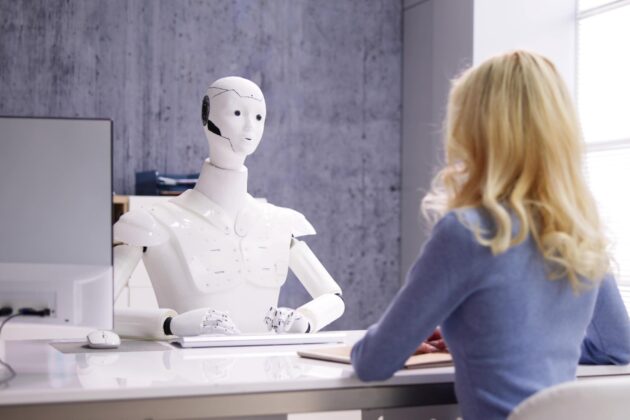 Robot Behind A Desk Conducting A Conversation With A Young Woman In Business Attire.